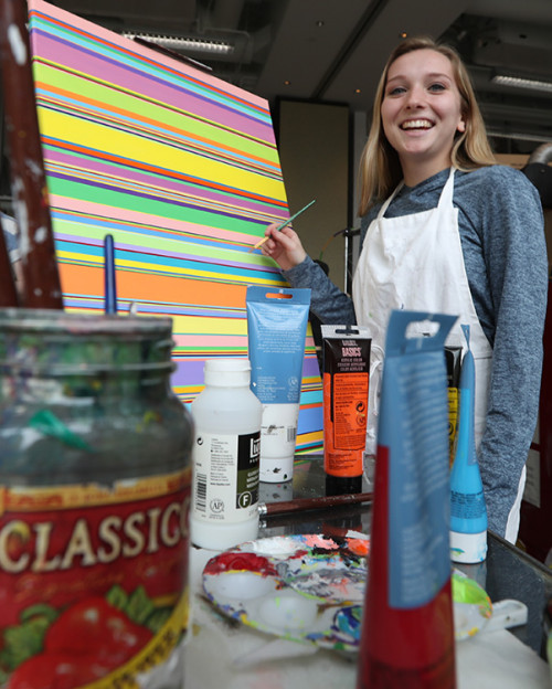 Female art student standing in front of her painting smiling. Her painting is horizontal lines of various widths and colors.
